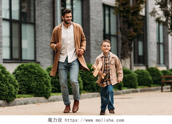 父亲和儿子在户外跑步爸爸在外套和儿子与玩具飞机在街上运行的全长视图